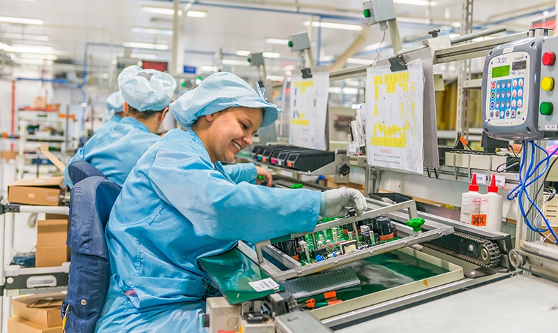 Smiling factory worker in a cleanroom environment assembling electronic components on a production line.