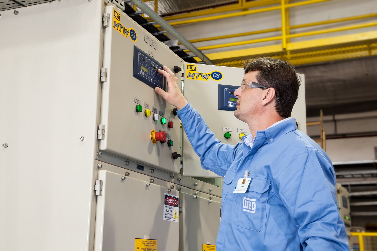 Technician operating an industrial motor control panel in a facility, wearing safety glasses and WEG-branded uniform, adjusting system settings.