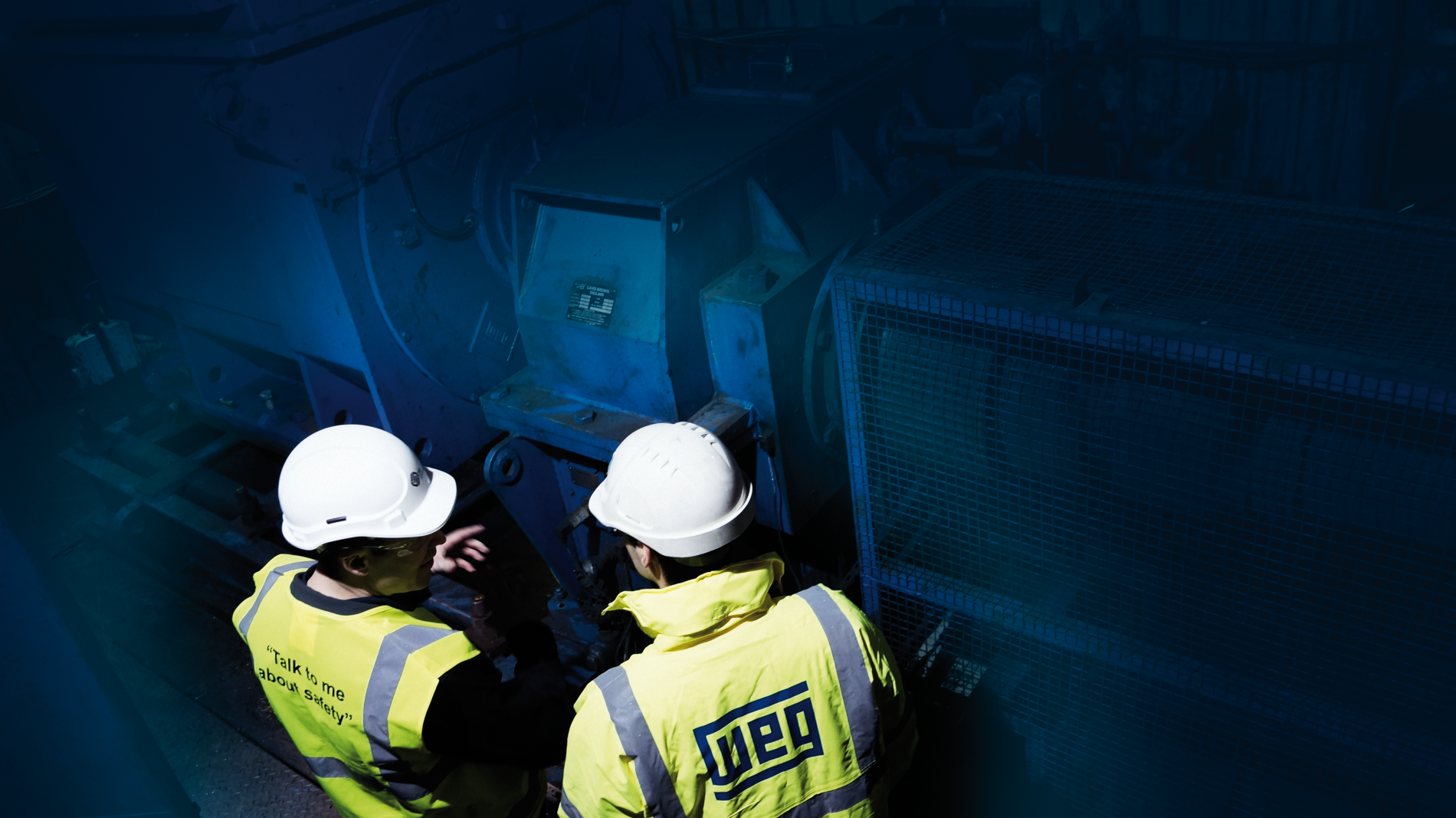 Two engineers in high‑visibility jackets and hard hats inspecting a large industrial motor inside a dimly lit facility.