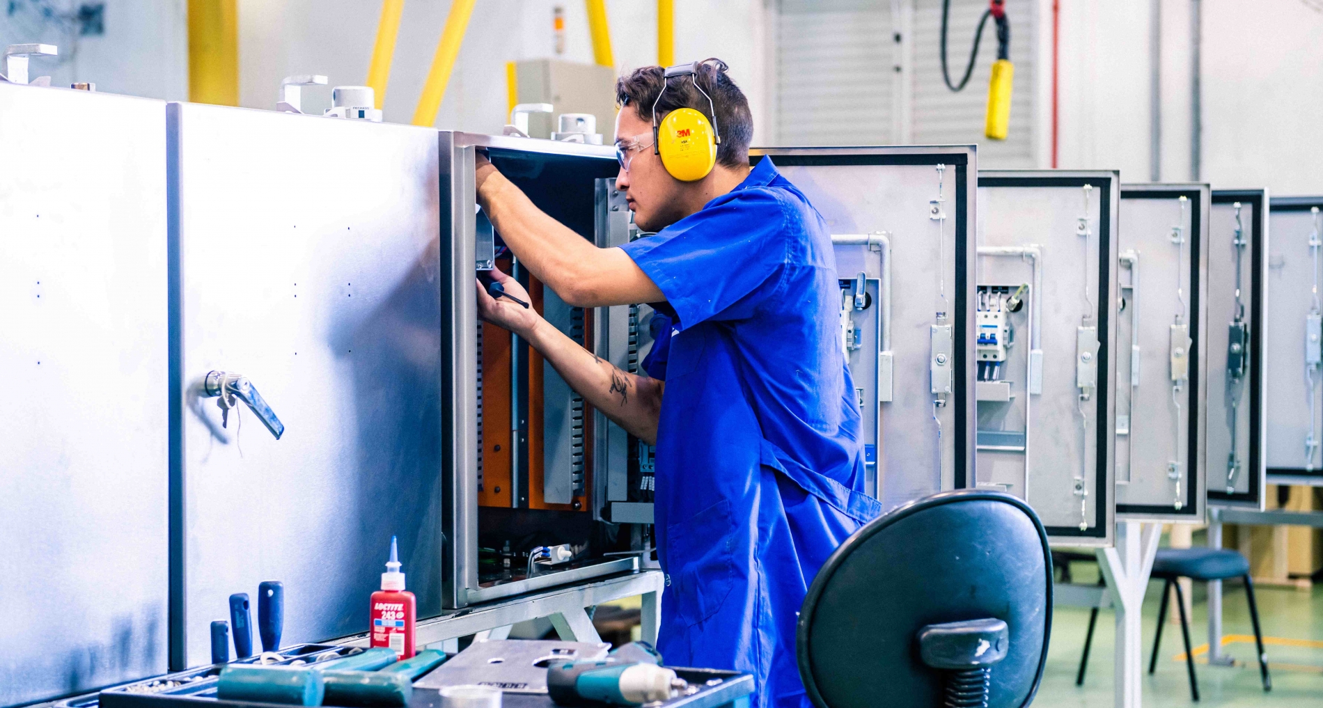 Technician in coveralls and ear protection wiring an industrial automation control panel in a workshop.