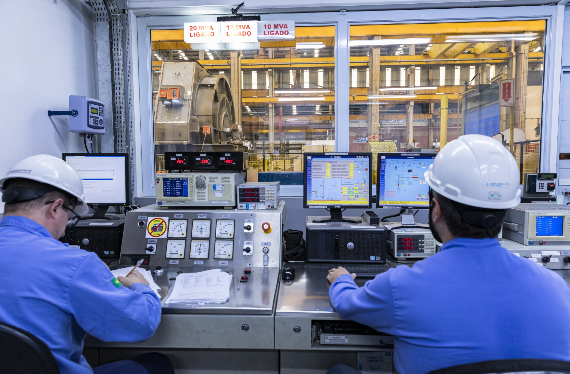Two technicians in an industrial control room monitor AC and DC motor systems on computer screens.