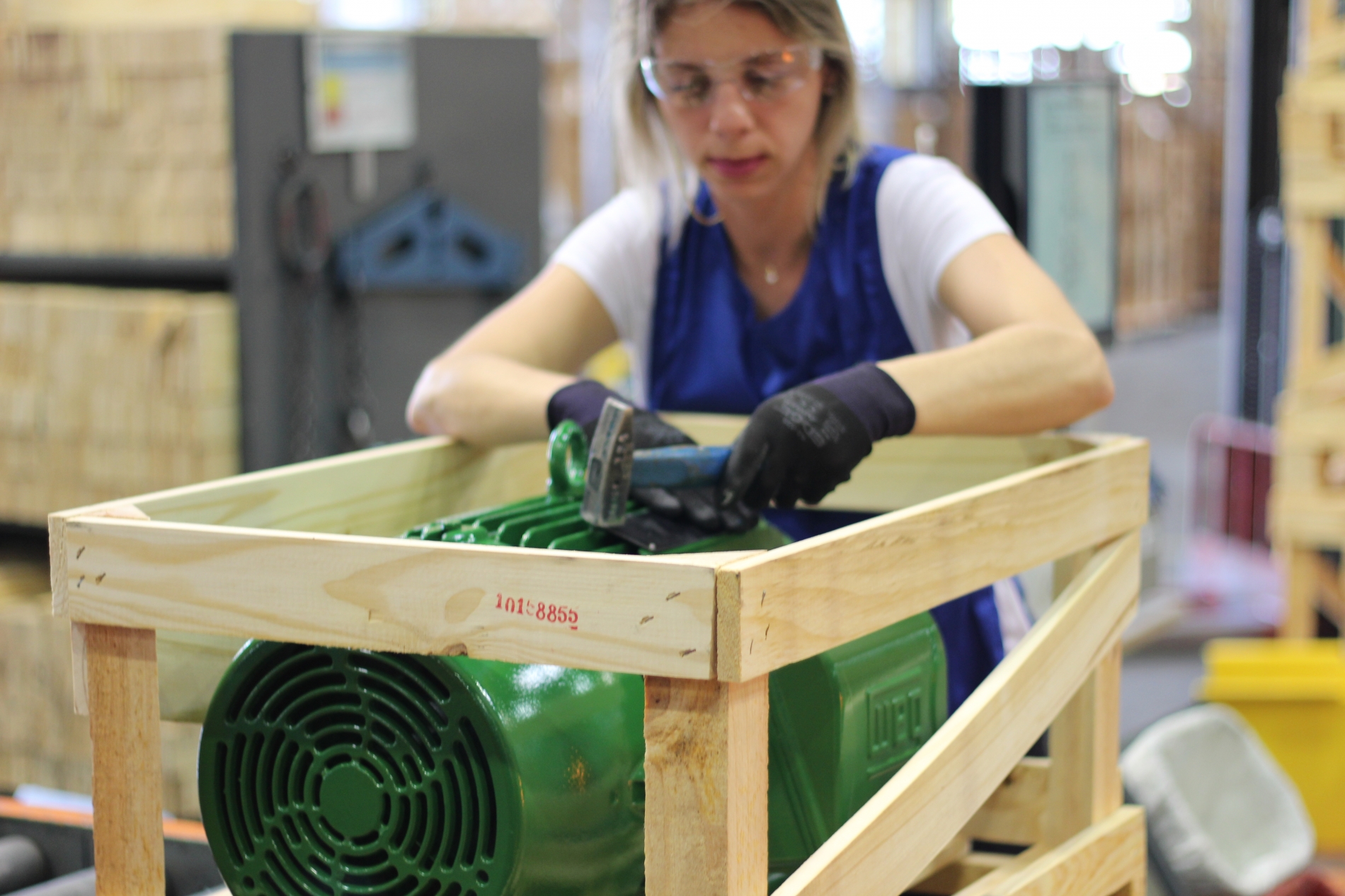 A technician wearing safety glasses and gloves secures a large green AC motor inside a wooden crate in an industrial warehouse.