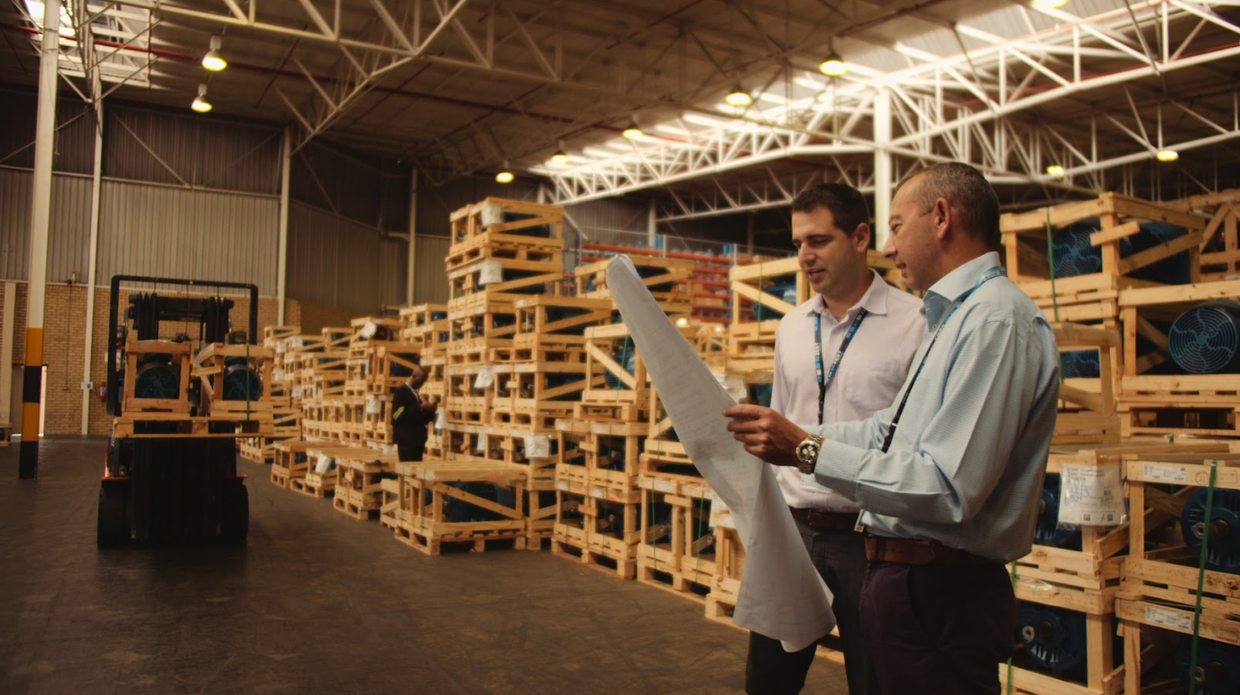 Two project managers review blueprints or documents in a well-lit warehouse filled with wooden crates of industrial equipment.