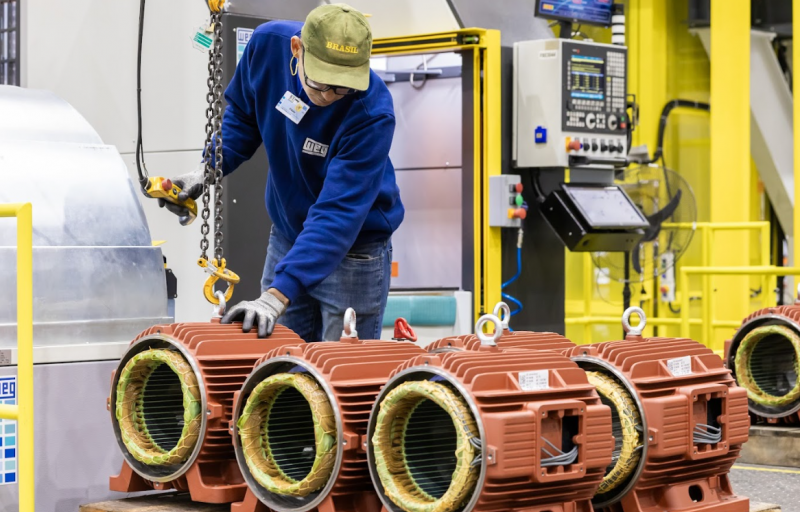 Technician lifting transformer parts with hoist equipment in a manufacturing facility.
